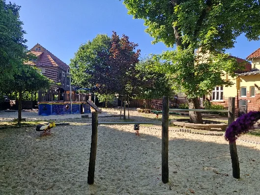 Das Außengelände der Marienschule: Sandfläche unter großen Bäumen, Wippfiguren, ein hölzernes Spielschiff und hinten das Gebäude.