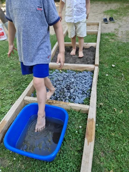 Kinder gehen barfuß über einen Fühlpfad im Garten, mit Feldern aus Erde, Steinen und einer Wanne mit Wasser.