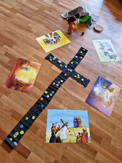 Bodenbild auf dem Holzfußboden: ein Kreuz aus schwarzem Stoff mit bunten Blüten, ringsum Bilder zur Ostergeschichte.