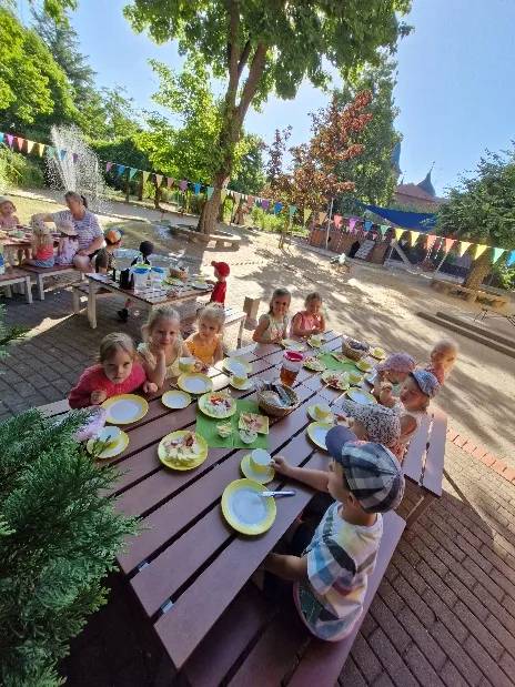 Kinder essen gemeinsam an einer langen Tafel im sonnigen Außengelände, darüber bunte Wimpelketten.