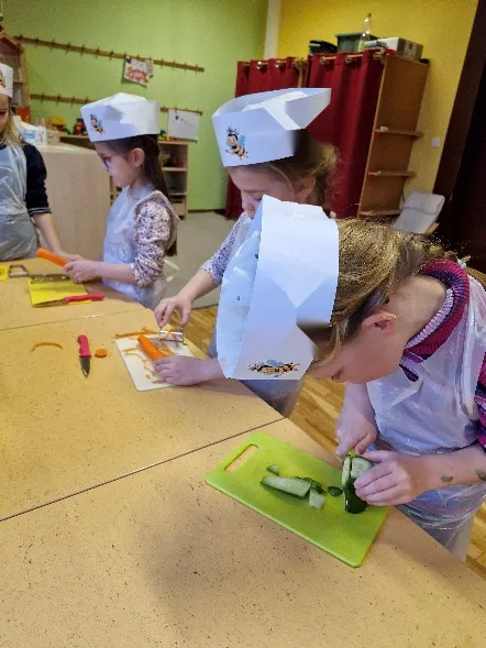 Kinder mit Papiermützen schneiden am Tisch Gurken und Möhren.