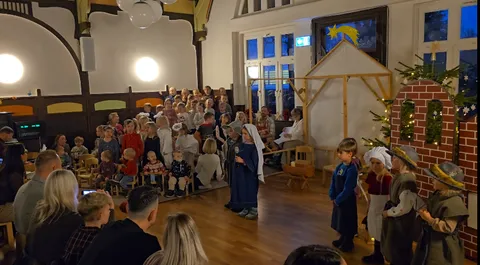 Krippenspiel im Saal: verkleidete Kinder als Maria, Josef und Hirten vor einem Stall aus Holz, Familien schauen zu.