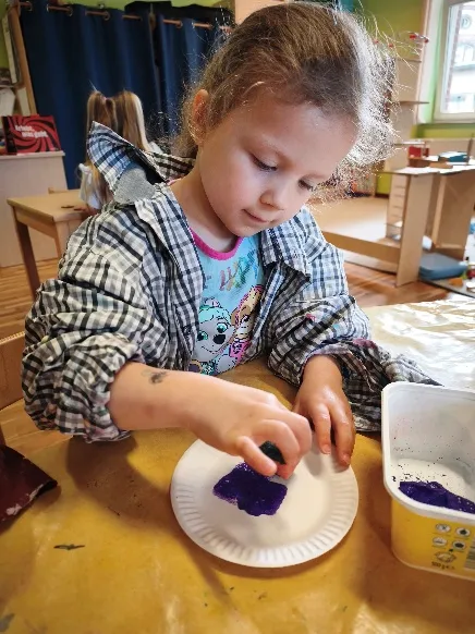 Ein Mädchen in einem Malerhemd tupft mit einem Schwamm violette Farbe auf einen Pappteller.