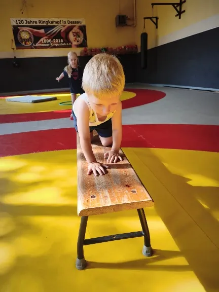 Ein kleiner Junge klettert in einer Sporthalle auf allen vieren über eine Holzbank.